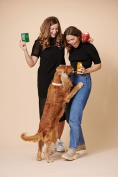 two women standing next to each other with a dog on their lap and one holding a drink in her hand