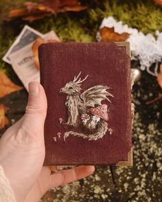 a hand holding up a small book with a dragon embroidered on it's cover