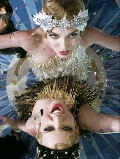 two women in costumes with their faces covered by sequins and feathers, looking up at the camera