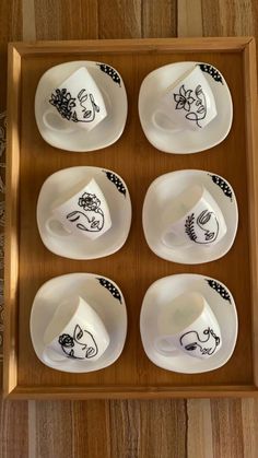 six white dishes with black designs on them in a wooden tray against a wood wall