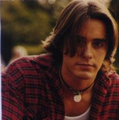 a young man with long hair wearing a red and black checkered shirt looking at the camera