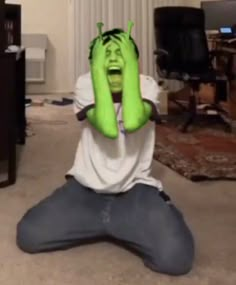 a man with green hands covering his face sitting on the floor in front of a tv