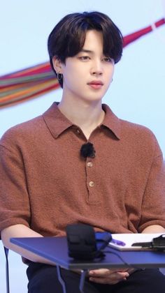 a young man sitting at a table with a microphone in front of him and wearing a brown shirt