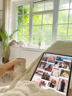 a woman sitting in bed holding a coffee cup and looking at her computer screen with photos on it