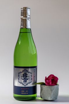 a bottle of wine next to a small vase with a pink flower in it on a white surface