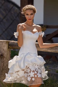 a woman in a short white dress leaning on a wooden fence with her hands behind her back