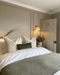 a bed with white sheets and pillows in a bedroom next to a door, lamp and potted plant