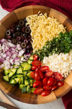 a wooden bowl filled with lots of different types of vegetables next to olives and cheese