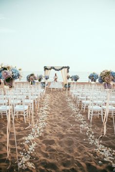 an outdoor ceremony set up with white chairs and blue flowers on the sand at the beach