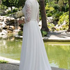 a woman standing in front of a pond wearing a white wedding dress with long sleeves