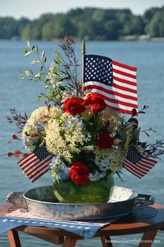 an american flag arrangement in a bucket on a table next to the water with red, white and blue flowers