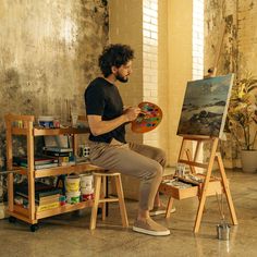 a man sitting in front of an easel holding a paintbrush and looking at the painting