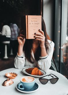 a woman sitting at a table with a book in front of her face and coffee