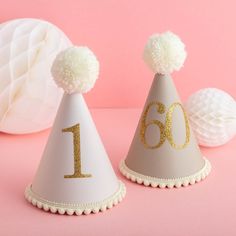 two white party hats with gold numbers and pom - poms on pink background