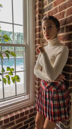 a woman in a plaid skirt leaning against a brick wall with her hands on her hips