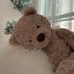 a brown teddy bear sitting on top of a white fluffy pillow next to a window