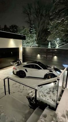 two cars are parked in the snow near a house at night with lights shining on them