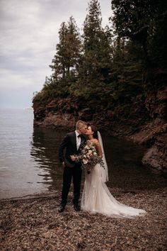 a bride and groom kissing on the shore of a lake with trees in the background