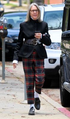 an older woman in plaid pants and black jacket walking down the street with her cell phone