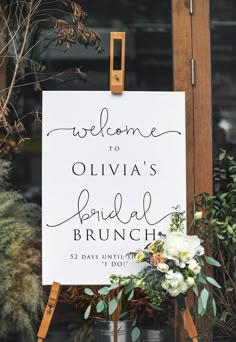 a welcome sign with flowers and greenery is displayed in front of a window for an outdoor bridal brunch