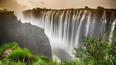 a large waterfall that is surrounded by trees and grass in the foreground, with water pouring over it