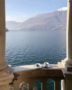 two tea cups sitting on top of a balcony next to the ocean with mountains in the background