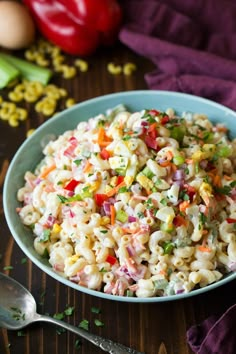 a blue bowl filled with macaroni salad on top of a wooden table next to vegetables