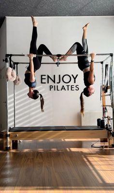 two women doing aerial acrobatic exercises in front of the enjoy pilates sign