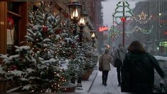 two people walking down a snowy street with christmas lights on the trees and buildings in the background