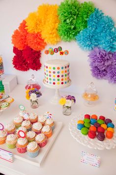 a table topped with lots of cupcakes next to a cake and rainbow decorations