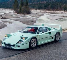 a light blue sports car driving down the road next to a river and mountains in the background