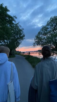 two people are walking down the road at dusk with trees and water in the background