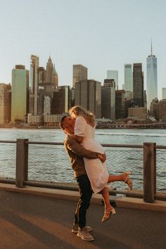 a man holding a woman on the side of a bridge over water with city buildings in the background