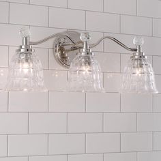 three light bathroom fixture with clear glass shades on white tile wall and subway tiles in the background