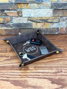 a leather tray with keys and wallets on top of a wooden table next to a stone wall