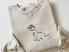 a white shirt with a mushroom on the front and a dinosaur embroidered on the back