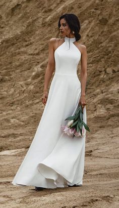 a woman in a white dress is holding flowers and walking down the beach with her hand on her hip