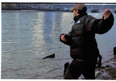 a man in black jacket standing next to water with birds on shore and bridge in background