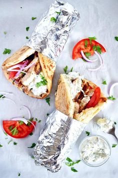 two pita sandwiches on foil with tomatoes and onions next to them, sitting on a white surface