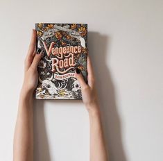 two hands holding up a book with an orange and black design on it, against a white wall