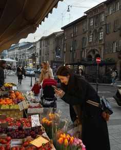 a woman looking at her cell phone while standing in front of a fruit and vegetable stand