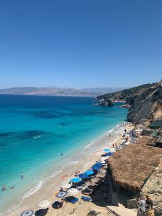 a beach with umbrellas and people on it next to the blue water in front of some buildings