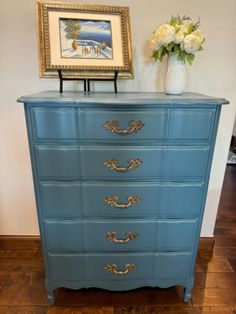 a blue chest of drawers with flowers on top and a framed painting above the drawer