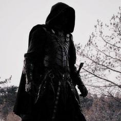 a man dressed in black standing next to a tree and looking at the sky with his hood up