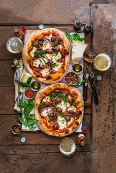 two pizzas sitting on top of a wooden table next to beer glasses and utensils