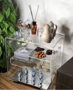 an acrylic bar cart with drinks and snacks on it in front of a potted plant