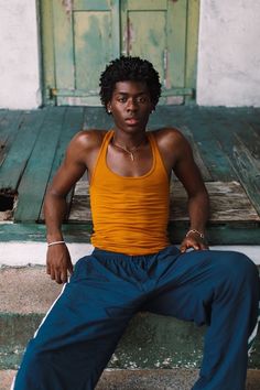 a young man sitting on the steps in front of a door wearing an orange tank top and blue pants