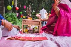 a woman sitting on the ground next to a bird cage with birds in it and people standing around