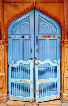 two blue doors are open in front of a yellow building with an arched door and iron bars