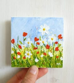 a hand holding up a piece of paper with flowers painted on it and grass in the background